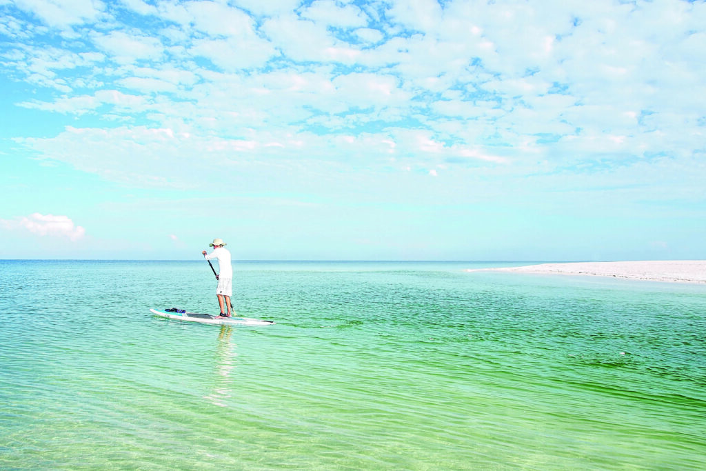 A man paddleboards on a clear summer day at Clam Pass where it meets the Gulf of Mexico.